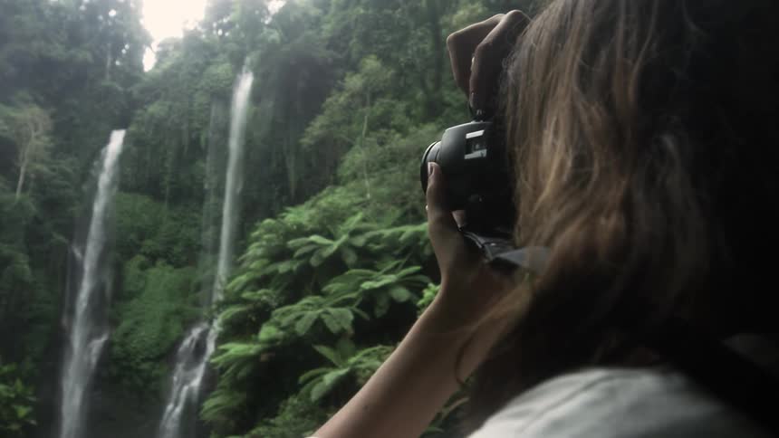 A woman is holding a camera and taking a picture of waterfalls in a tropical jungle