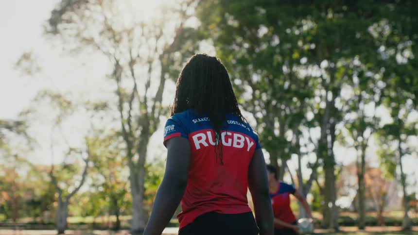 Girls wearing jerseys are throwing a rugby ball to each other in a park