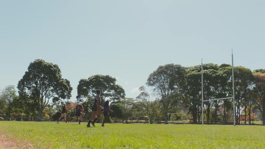 A group of female rugby players toss a rugby ball while running through an athletics field during the day