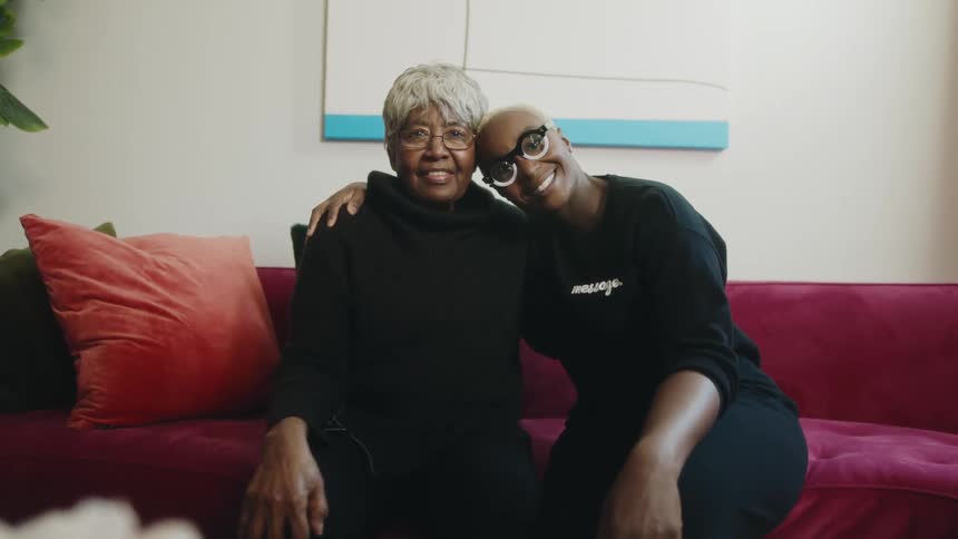 A woman hugs her elderly mother as they smile into the camera while sitting on a sofa in the living room of a house