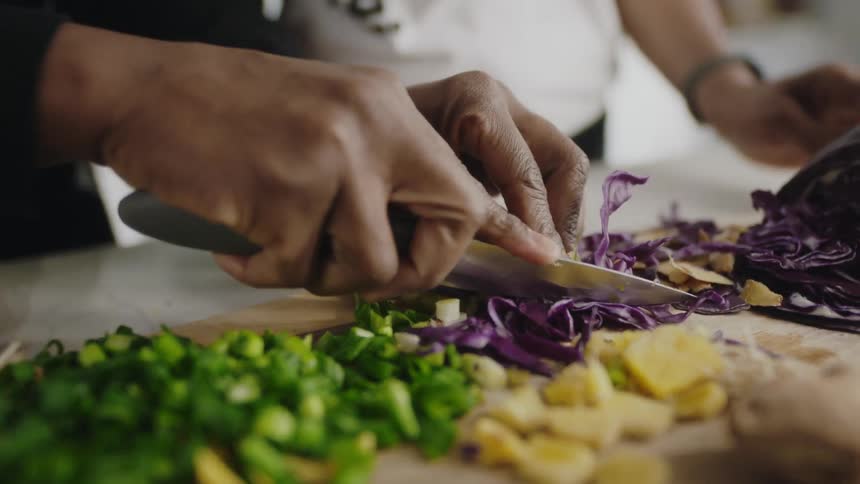 One person carefully slices shredded cabbage on a wooden cutting board as another person looks on