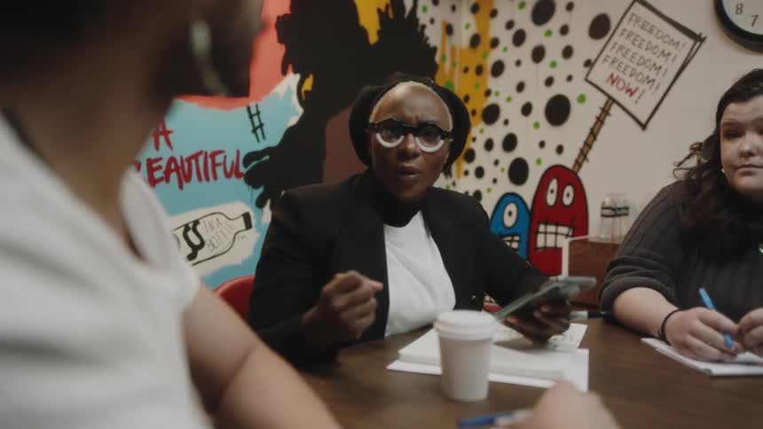 A woman gestures as she talks and leads a meeting in a room with activism mural on the wall