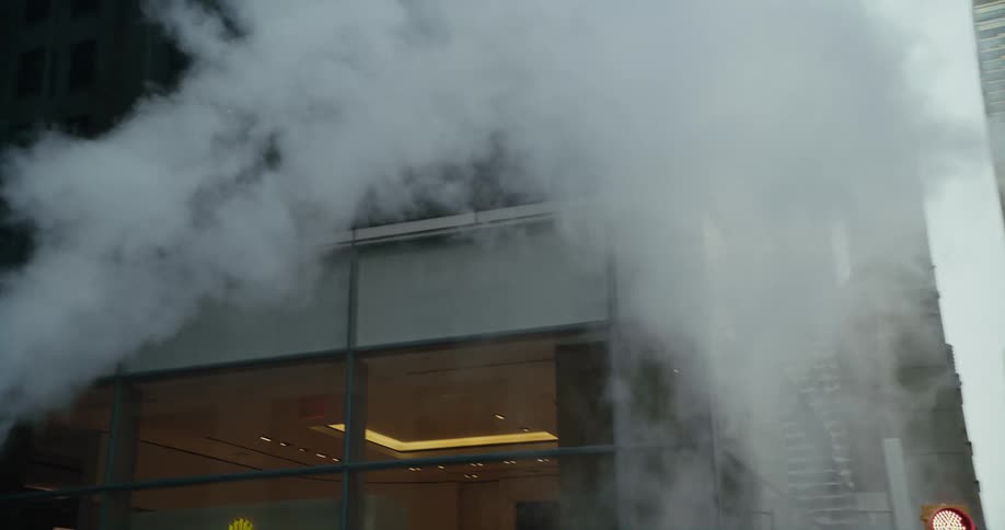 Steam is coming out of a pipe on a city street and rising around a building