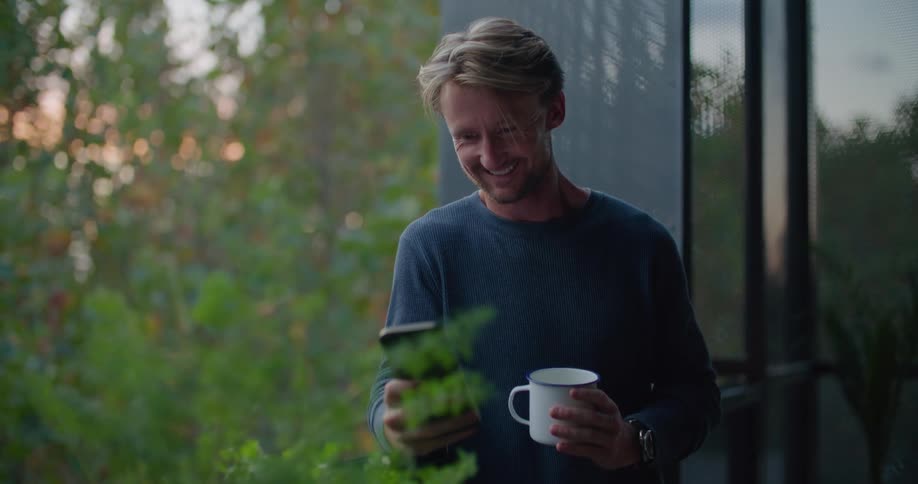 A man leaning against his balcony railing texting on a phone smiling and holding a mug