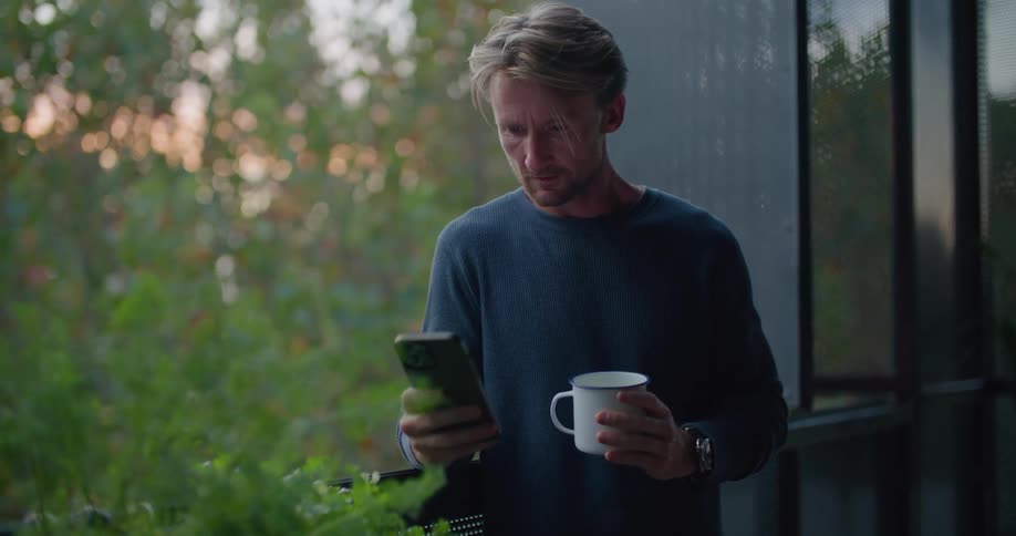Young man reclining on a window drinks from a white cup while texting on the phone at sunset