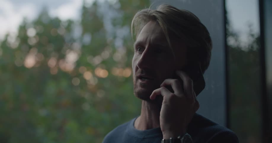 A man stands next to a large window while talking into his cell phone