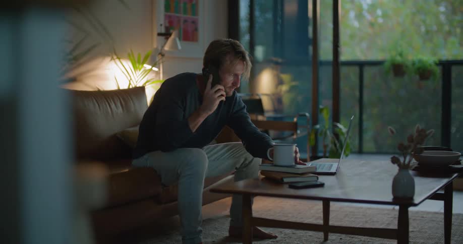 A man sits down and uses a cell phone and laptop