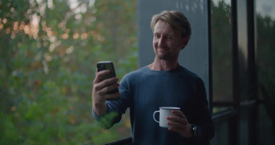 A man is standing outside on a balcony holding a coffee cup and a cell phone and he says something when he looks at the phone