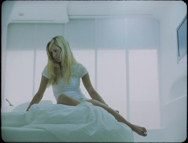 A woman dressed in white sits on a white bed in a white room with the white window shade rising behind her