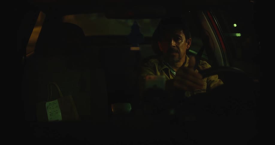 A man is driving an automobile at night and there is a paper bag in the passenger seat as he talks to someone unseen