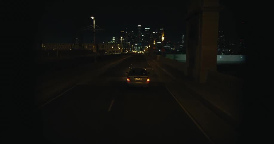 A car drives along a bridge in the dark of night as it approaches a city