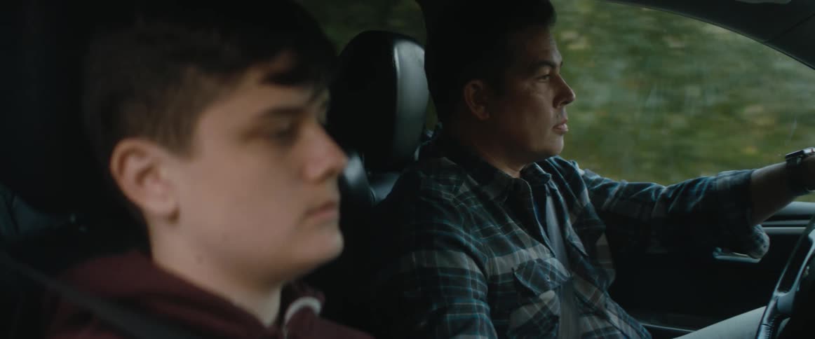 A man is driving an automobile and a teenage boy is in the passenger seat