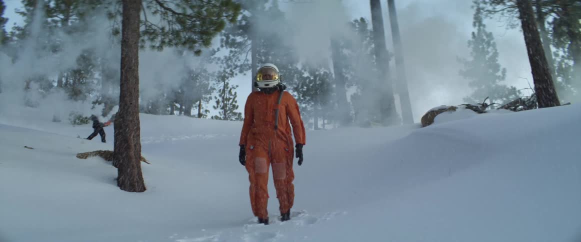 Person wearing pilot crash suit walks down snow covered slope in conifer forest as man spreads smoke in the background