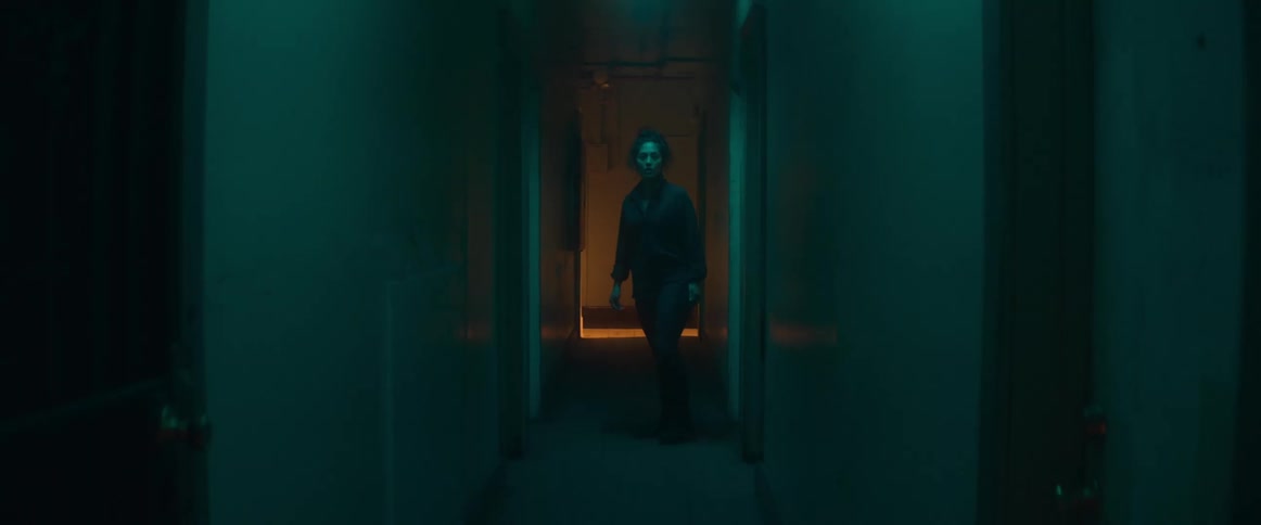 A woman looks down a dimly lit hallway in an apartment building and walks through a doorway