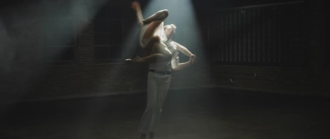 Male dancer with white shirt and pants dances with female dancer wearing white dress in spotlight