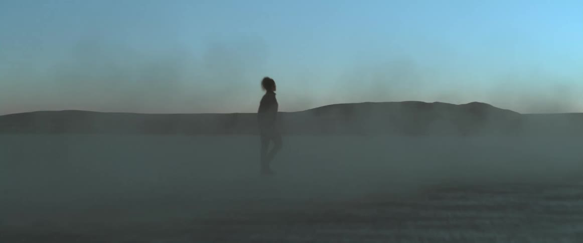 A woman is walking in the desert as a truck with headlights on passes her and there are mountains in the distance