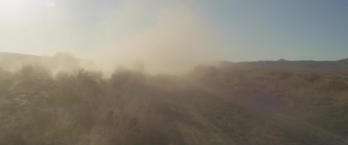 The sun shines brightly upon a truck traveling on a rural dirt road and there are mountains in the distance