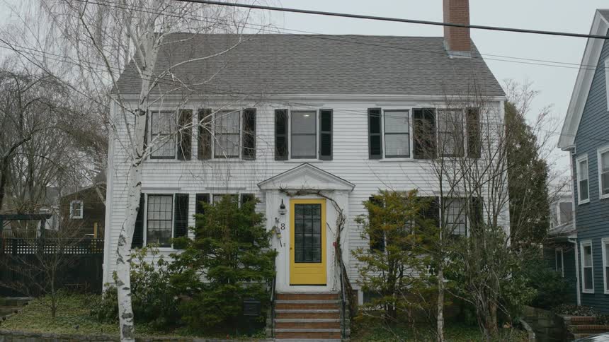 A white Colonial style house with brown shutters and a yellow door with steps leading up to it sits in a quiet neighborhood with bare birch trees and evergreen shrubs in front of the house.