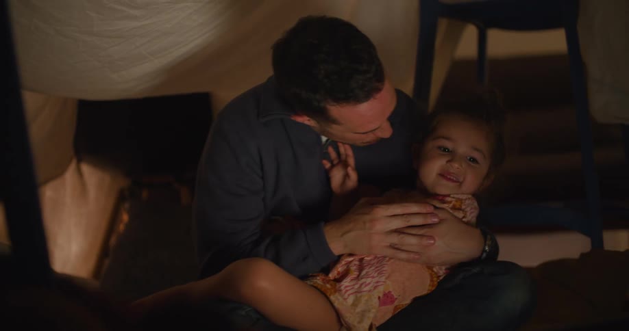 Father holds daughter in his lap and they smile and hug as they sit under impromptu sheet tent with light from screen shining onto them