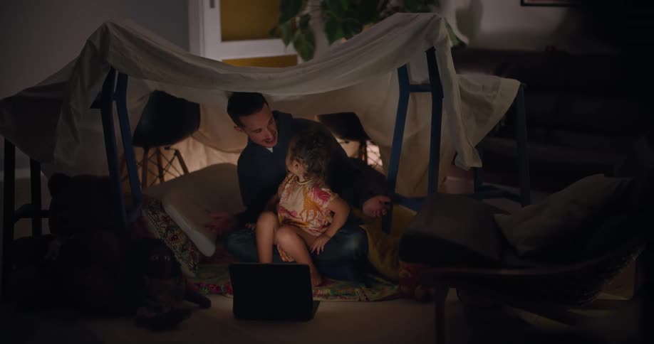 Father sits with daughter in his lap under impromptu sheet tent in home as they watch laptop