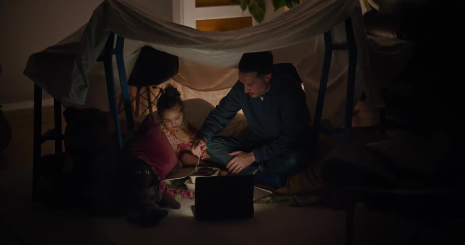 A dad sitting inside a sheet fort with his daughter trying to get her to eat takeout food