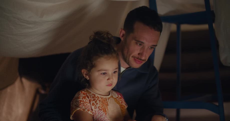 A man is sitting on the floor with his young daughter in his lap and a laptop computer in front of them