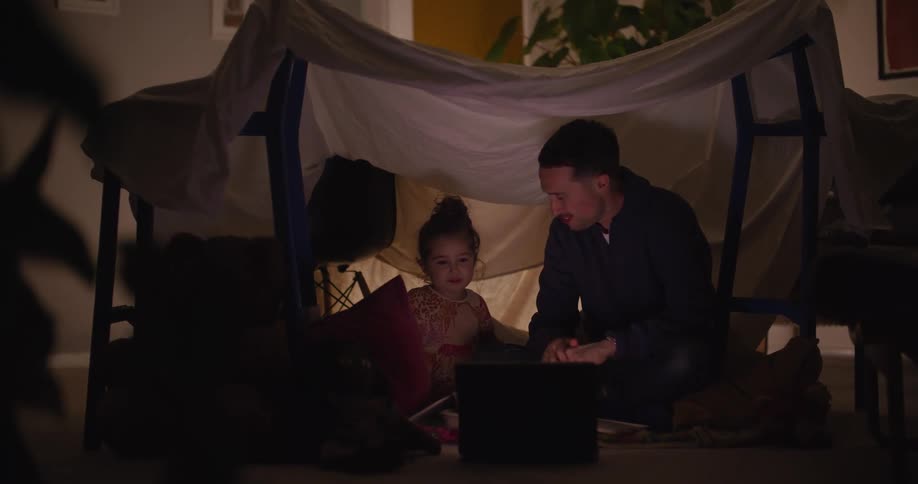Father and daughter sit on floor in home under an impromptu sheet tent eating and watching a show on a laptop