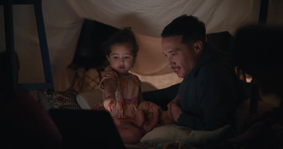 Father and daughter sit under impromptu sheet tent in home watching laptop