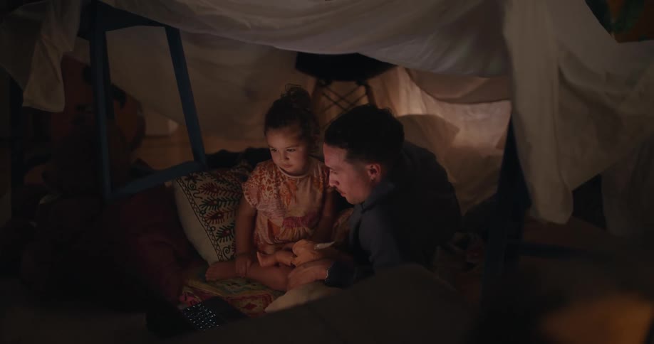 A father and his daughter laugh as they sit in a tent and watch something on a laptop