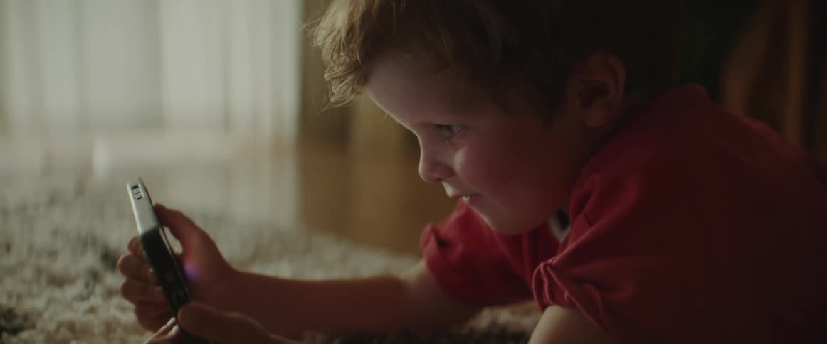 A young boy is lying on a rug on the floor holding a cell phone and looking at it