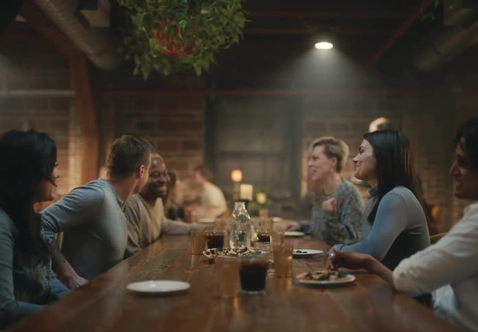A group of men and women laugh and talk and sit a table in a restaurant with food and drinks and another man comes and sits down and joins them