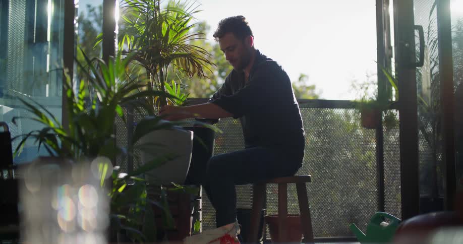 A man transfers soil into a flowerpot containing a large plant while sitting on a stool on a balcony