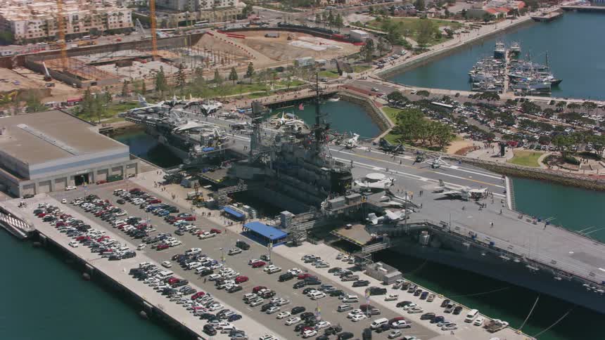Airplanes are on an aircraft carrier docked along the coast and there are cars in a parking lot on shore and boats in the marina