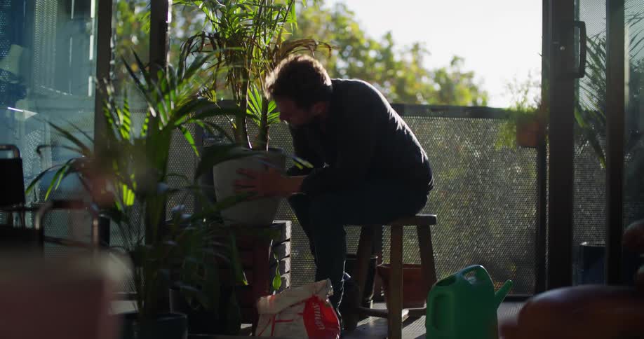 A man is sitting outside on a stool watering plants in pots