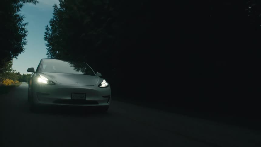 A white automobile is traveling on a rural road and moves into a forest area and the headlights shine in the darkness