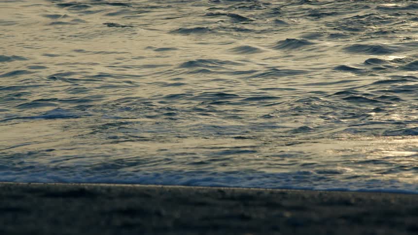 Sunlight reflects on the water as waves roll into shore and over the sand on a beach