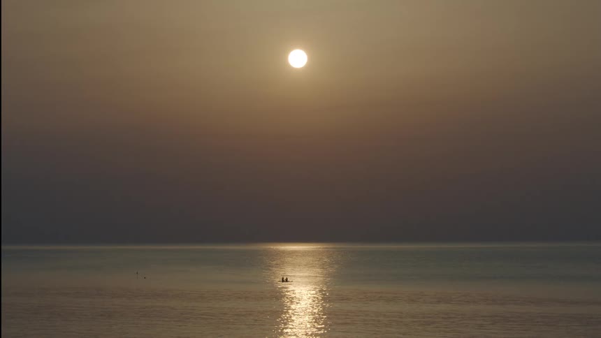 Silhouettes of two people in a canoe as the sun in a hazy red sky reflects on the water