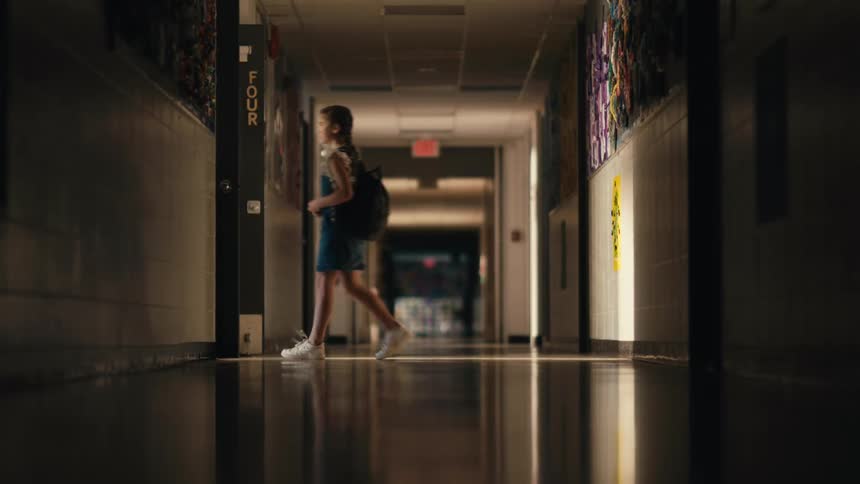 Teacher stands from kneeling to greet girl wearing backpack then leads girl into lit room