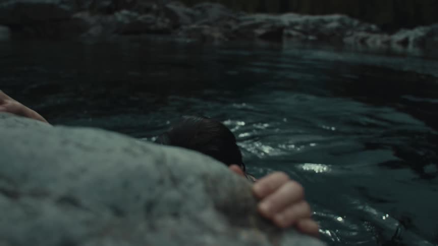 Smiling woman swimming in lake clings onto stone shore with forest in the background