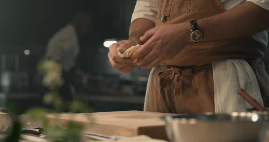 A cook kneads dough with his hands as another cook prepares food on a counter across the kitchen