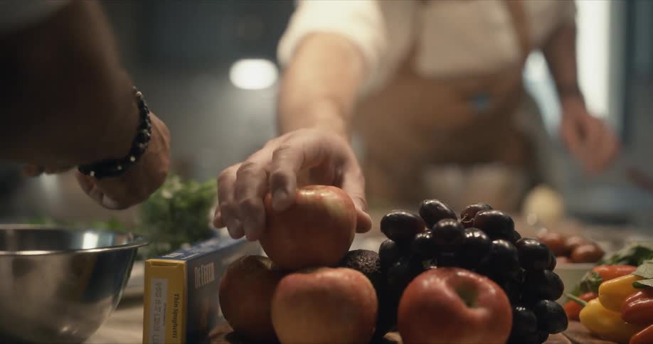 A man reaches across the counter for an apple while sharing the kitchen with another chef