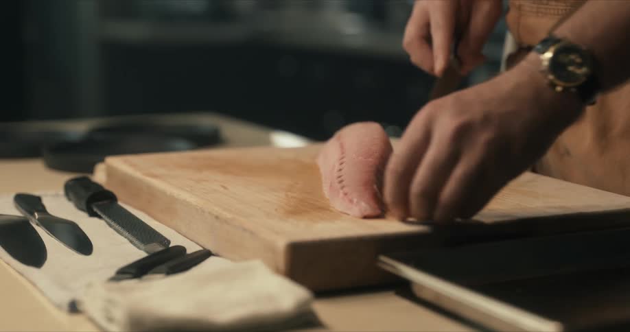 A man moves raw meat from a pan onto a wooden cutting board and begins to slice it with a knife