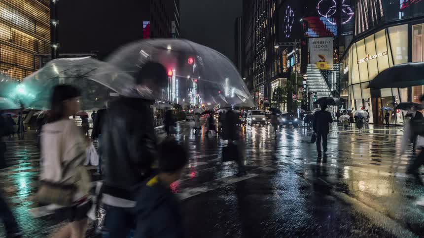 People are walking on a downtown street at night in the rain carrying umbrellas and lights from cars reflect on the road