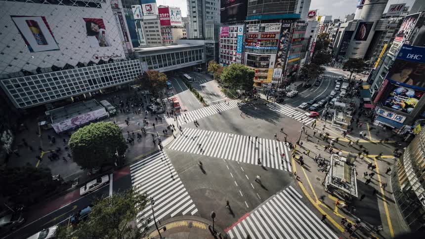 Pedestrians and traffic flows through busy intersection bordered by building with media screen
