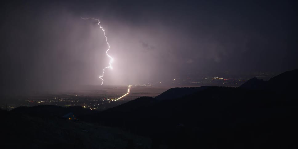 Lights are on in a house on a hill and a valley below is lit up as lightning flashes in the night sky above