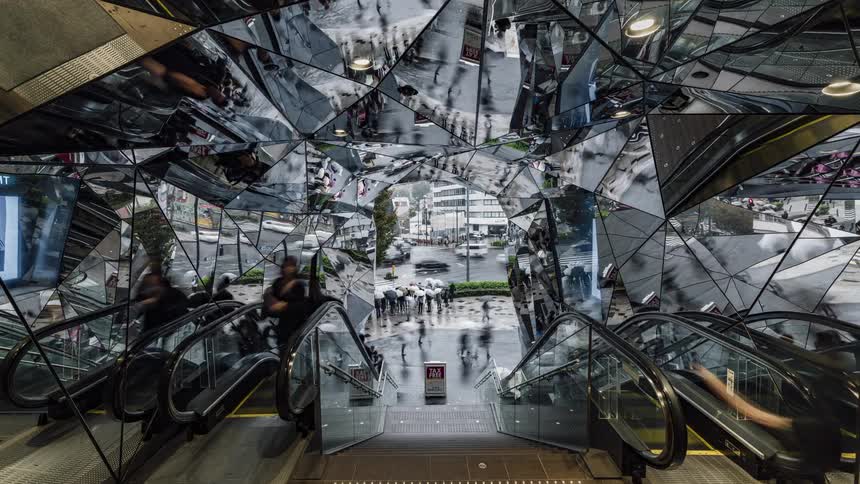 People are moving up and down an escalator as their reflections are on the mirror walls and ceiling and traffic is moving on the city streets outside