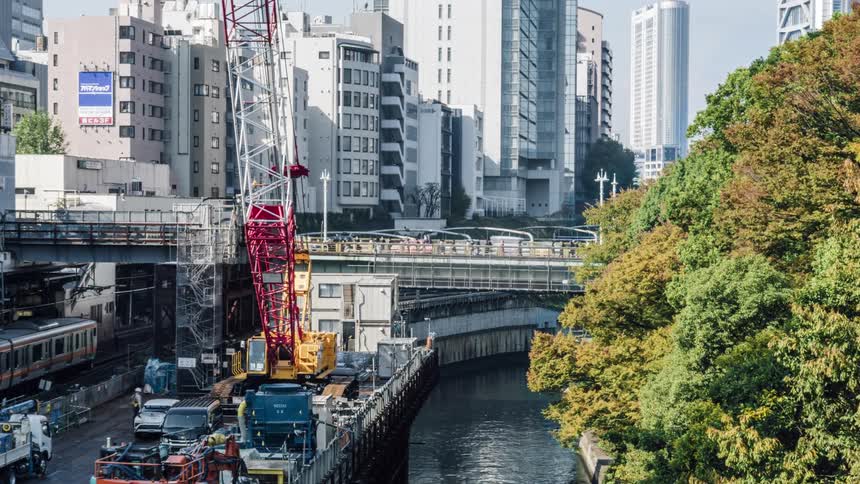 Pedestrians and automobiles cross a bridge over a river as a crane sits on the dock below and tall city buildings are in the distance