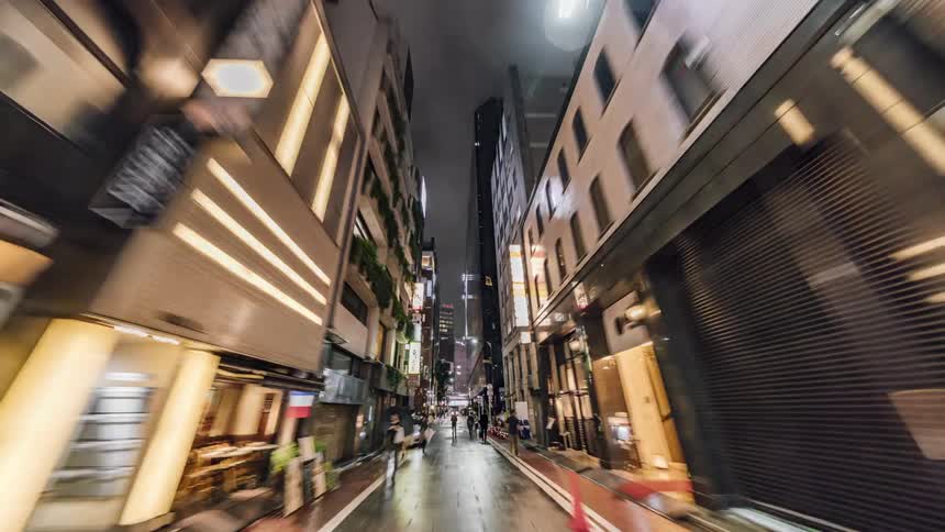 People are walking on a downtown street at night in the rain and lights reflect on the wet road