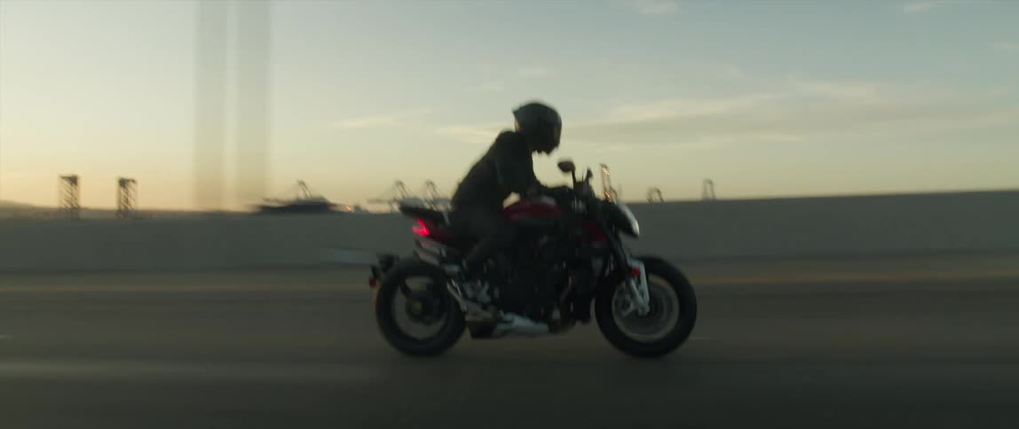 A cyclist wearing a black helmet is riding a motorcycle on a highway passing trucks and there are utility towers in the distance