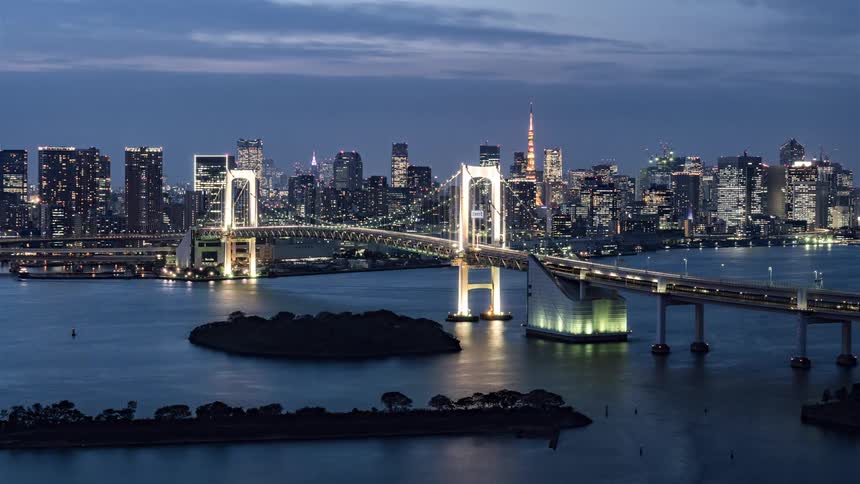 Traffic moves over a bridge leading to a big city as boats sail on the water below and the city lights go on when it gets dark outside